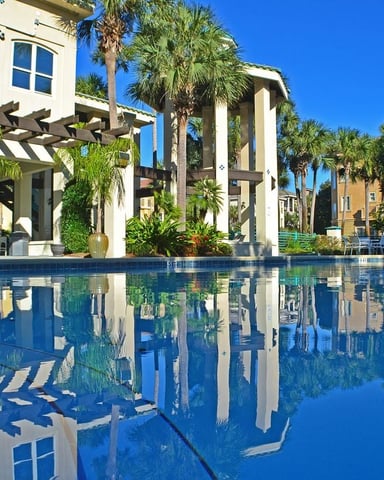 Resort pool with white pergola, palm trees, and multi-story buildings reflected in clear blue water