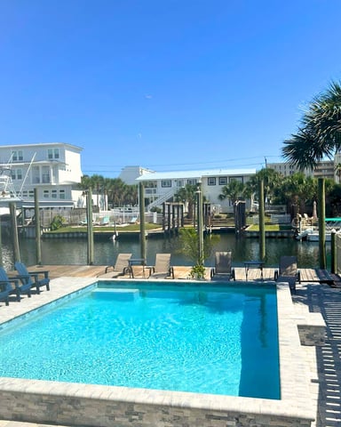 Waterfront pool with blue loungers, dock, palm trees, and white residential buildings under clear sky